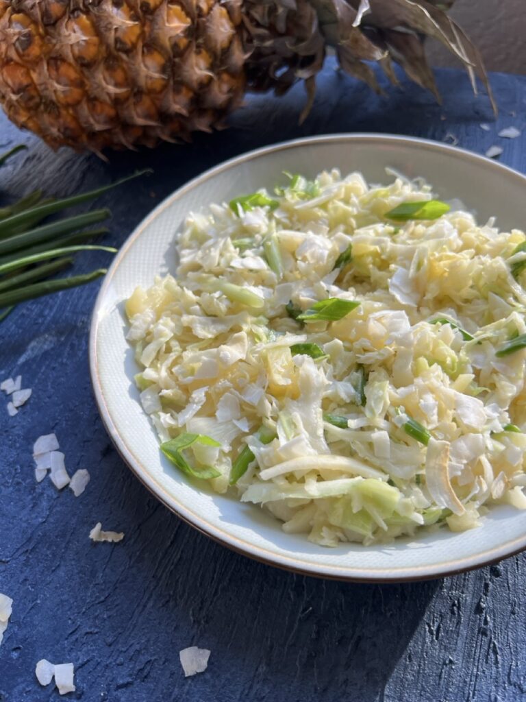 Bowl of tropical coleslaw with shredded coconut, crushed pineapple, and scallions with a fresh pineapple in the background.